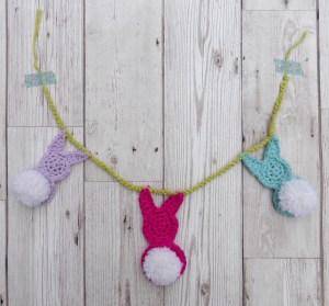 grey wooden floor background, crochet bunting with 3 rabbit shapes in purple, pink and blue with white pompom tails