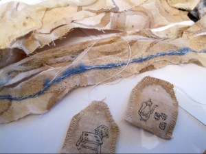 scraps of light brown fabric behind 3 fabric tea bags with images of tea pot and cups on one, and 2 stools on the other