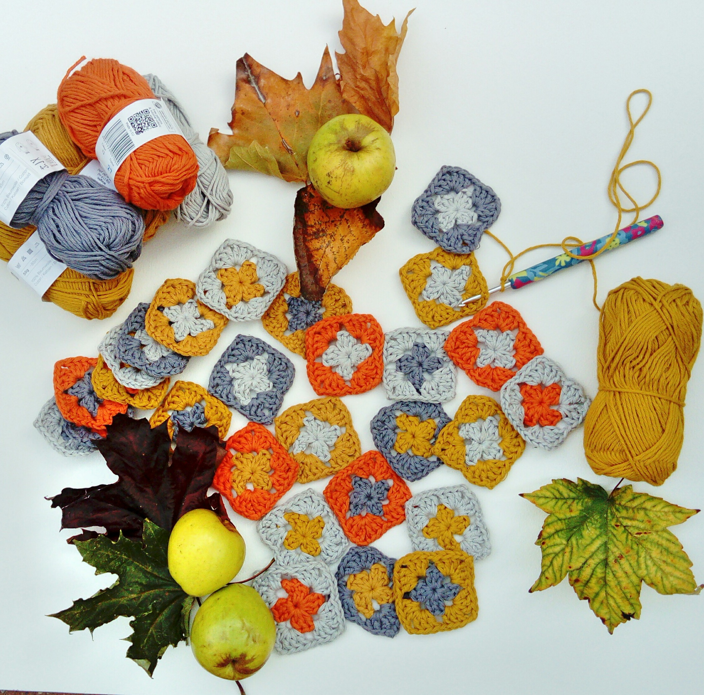 crochet squares in orange, grey, and yellow, surrounded by leaves and green apples