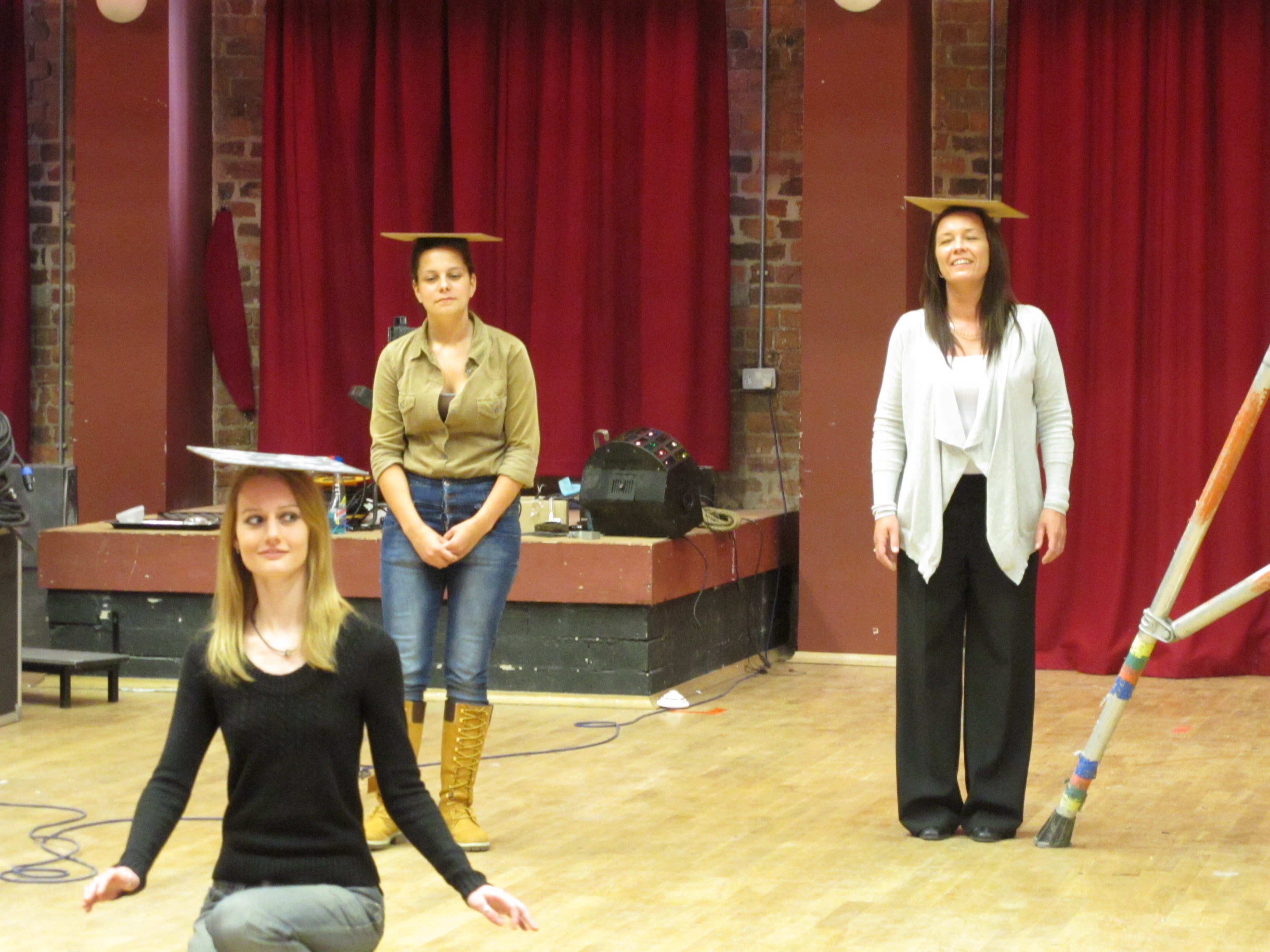 3 women with square boards on their heads. The woman in the foreground is kneeling trying to keep balance, the other 2 are stood up smiling