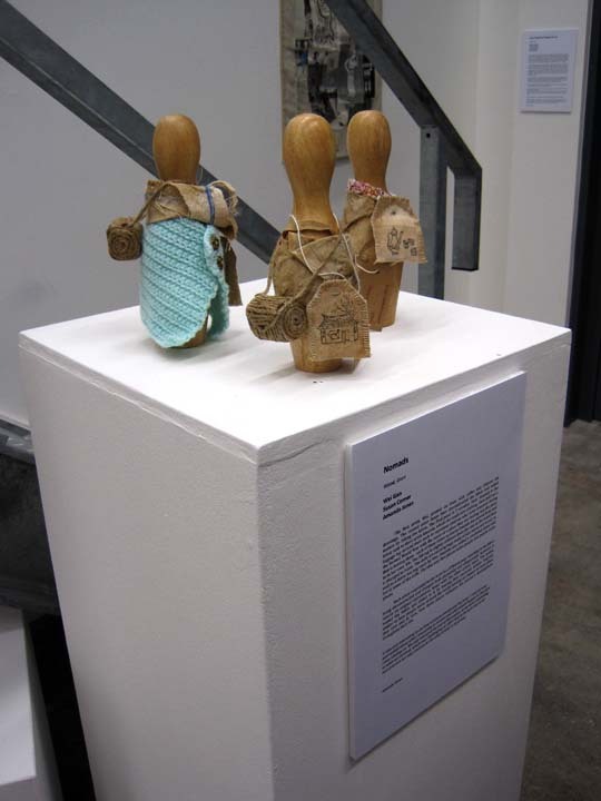 3 wooden dolls with no human features, dressed in tea stained material on a white plinth in a gallery