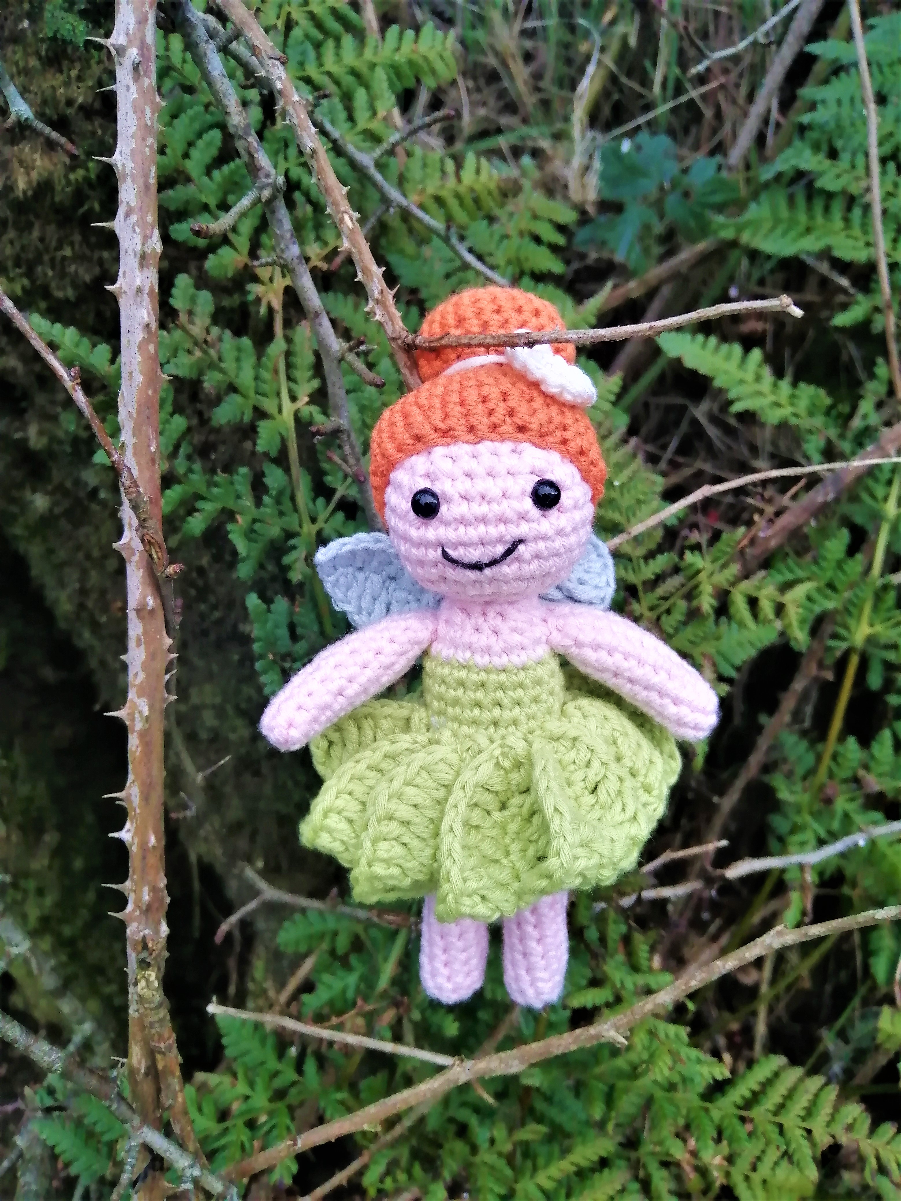 crochet pink skinned fairy with orange hair in a bun, green dress, with skirt part  looking like leaves. light blue wings. she has a smiling face and is stood among woodland trees. 